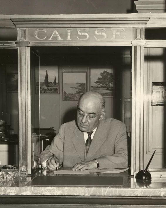 Black and white photograph of a bank employee in a suit, behind a counter marked ‘Cashier’.